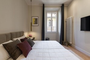 Un dormitorio con cama, mesita de noche, radiador, ventana y televisor montado en la pared.