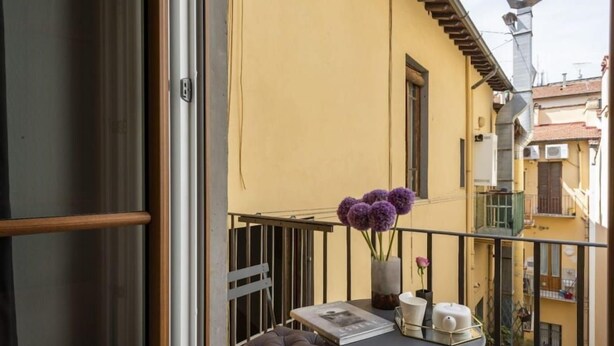 Ein Balkon mit Tisch, Kissen, einer Blumenvase und einem Fenster.