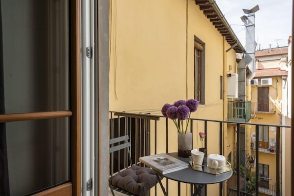 Un balcón con una mesa, cojines, un jarrón con flores y una ventana.