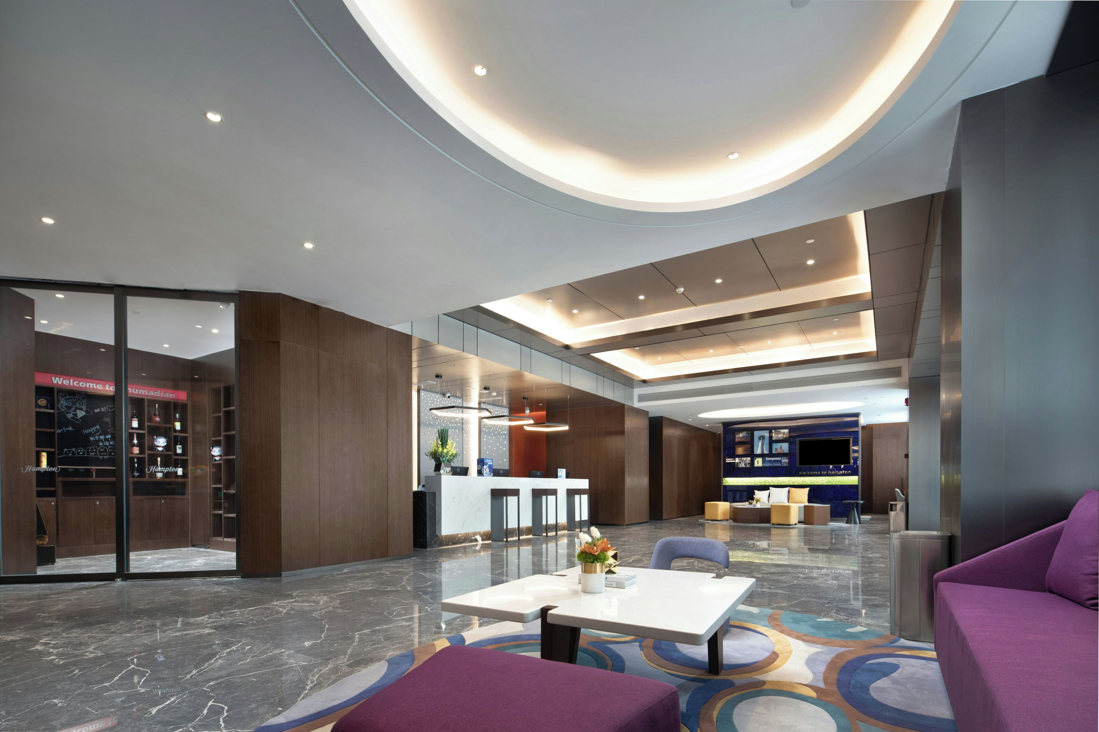 A modern hotel lobby with a reception desk, seating area, and a large screen.