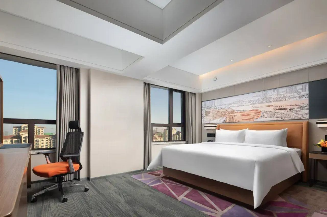 A hotel room with a large bed, a desk with a chair, a cityscape view, and a painting on the wall.