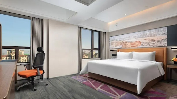 Una habitación de hotel con una cama grande, un escritorio con silla, vista panorámica de la ciudad y un cuadro en la pared.