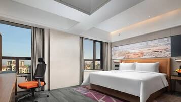 A hotel room with a large bed, a desk with a chair, a cityscape view, and a painting on the wall.