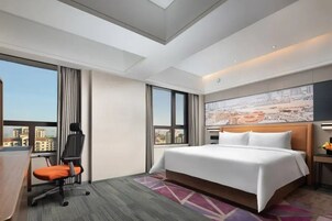 A hotel room with a large bed, a desk with a chair, a cityscape view, and a painting on the wall.