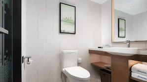 Un baño moderno con inodoro, lavamanos y espejo. Hay cuadros enmarcados en la pared y una cesta debajo del lavamanos.