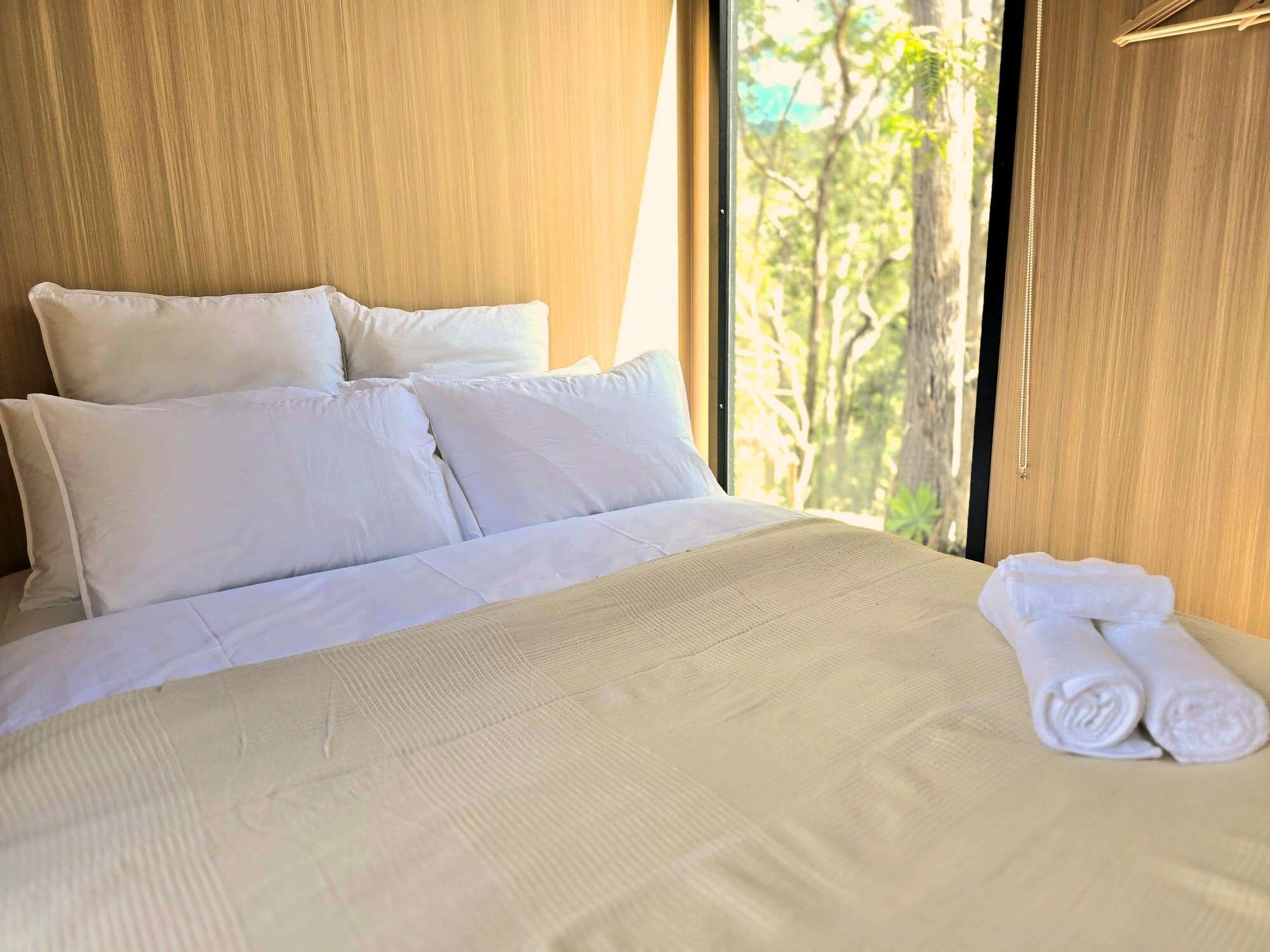 A neatly made bed with white linens and pillows, a pair of rolled towels, and a view of trees through a window.