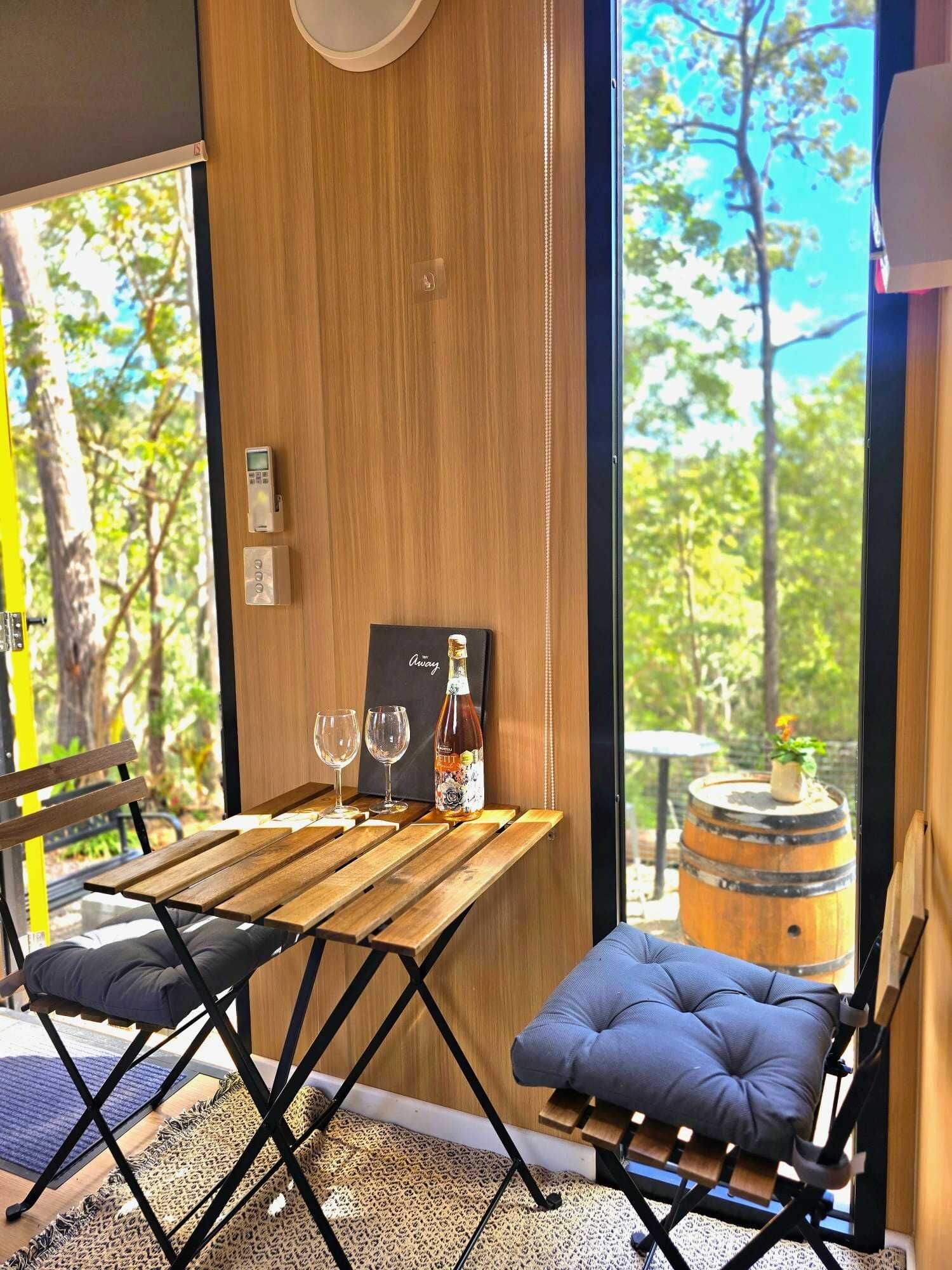 A wooden table with two wine glasses and a bottle, a blue cushioned chair, and a view of trees through large windows.