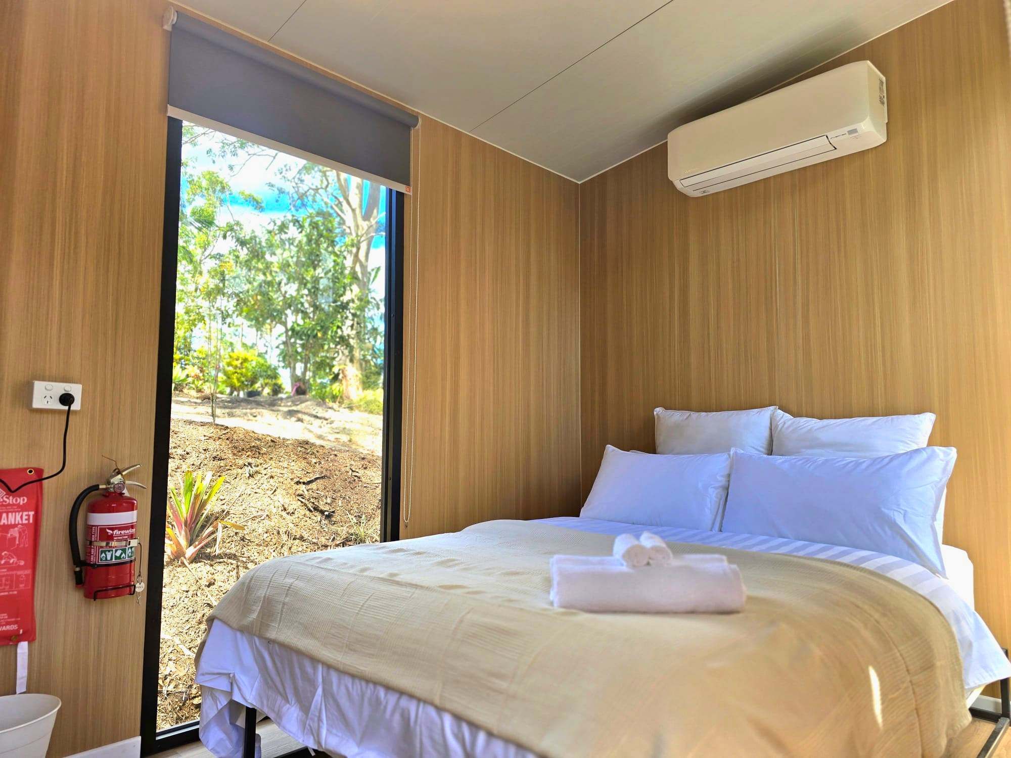 Ein Hotelzimmer mit einem Bett, weißer Bettwäsche, einem Feuerlöscher, einer Klimaanlage und Blick ins Freie.