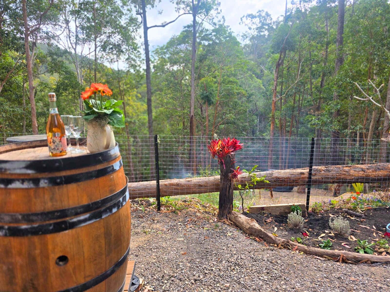 Rustikale Outdoor-Szene mit einem Holfass, einer Flasche und einem Glas auf einem Tisch, umgeben von einem Zaun und einem Wald im Hintergrund.