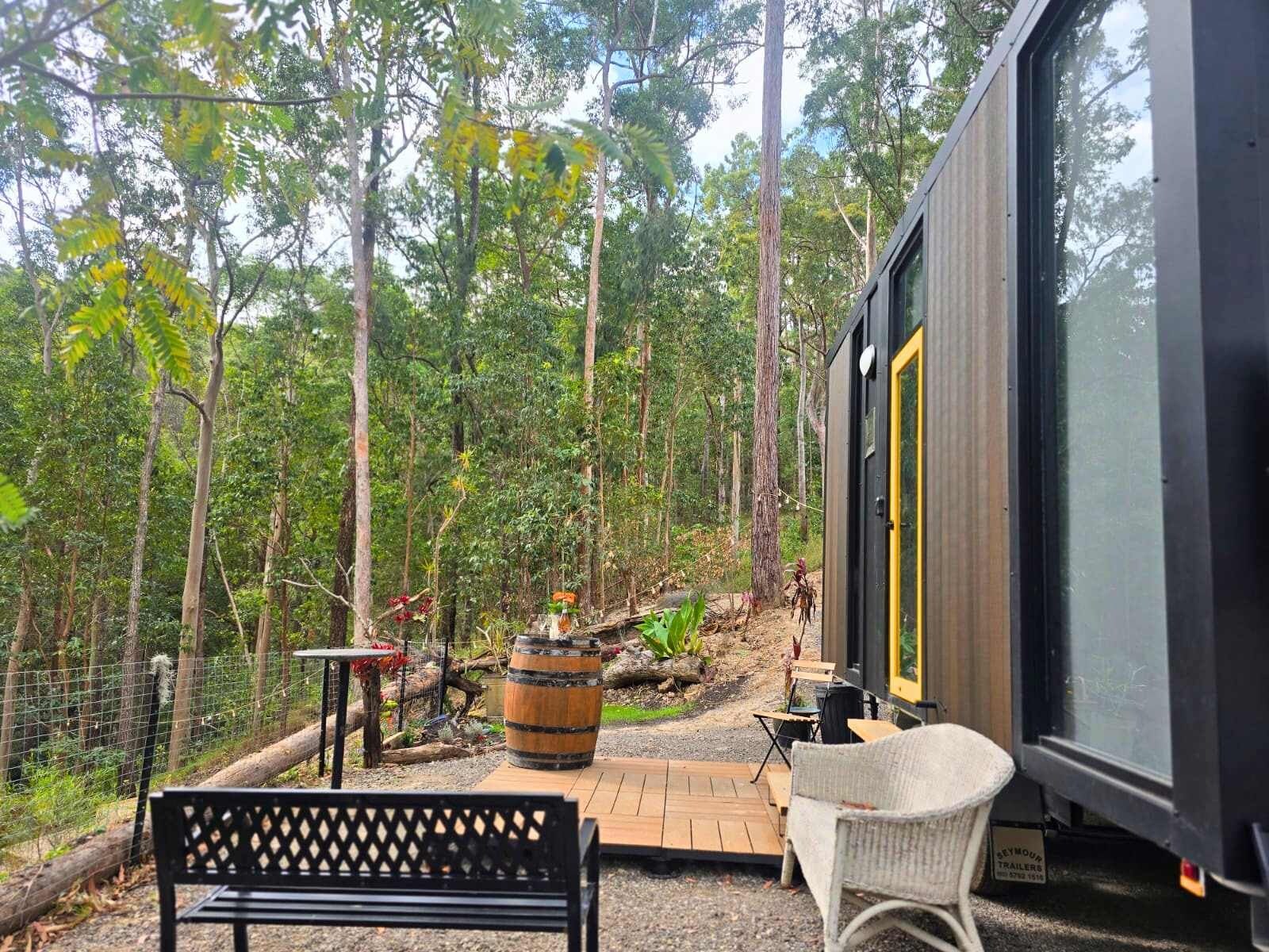 A modern cabin with a large window, a bench, and a rustic outdoor area with a barrel and a table.