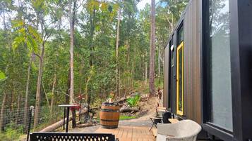 A modern cabin with a large window, a bench, and a rustic outdoor area with a barrel and a table.