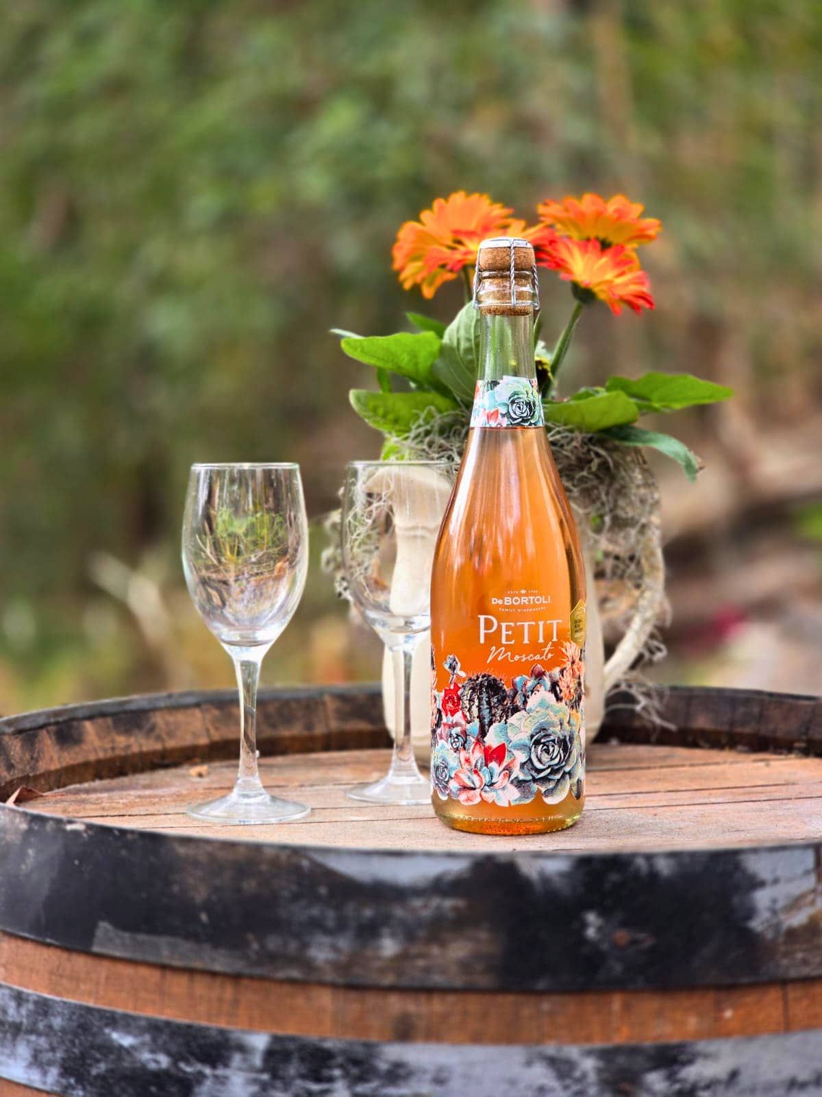 A bottle of Petit's wine on a wooden barrel with a glass and a vase of flowers.