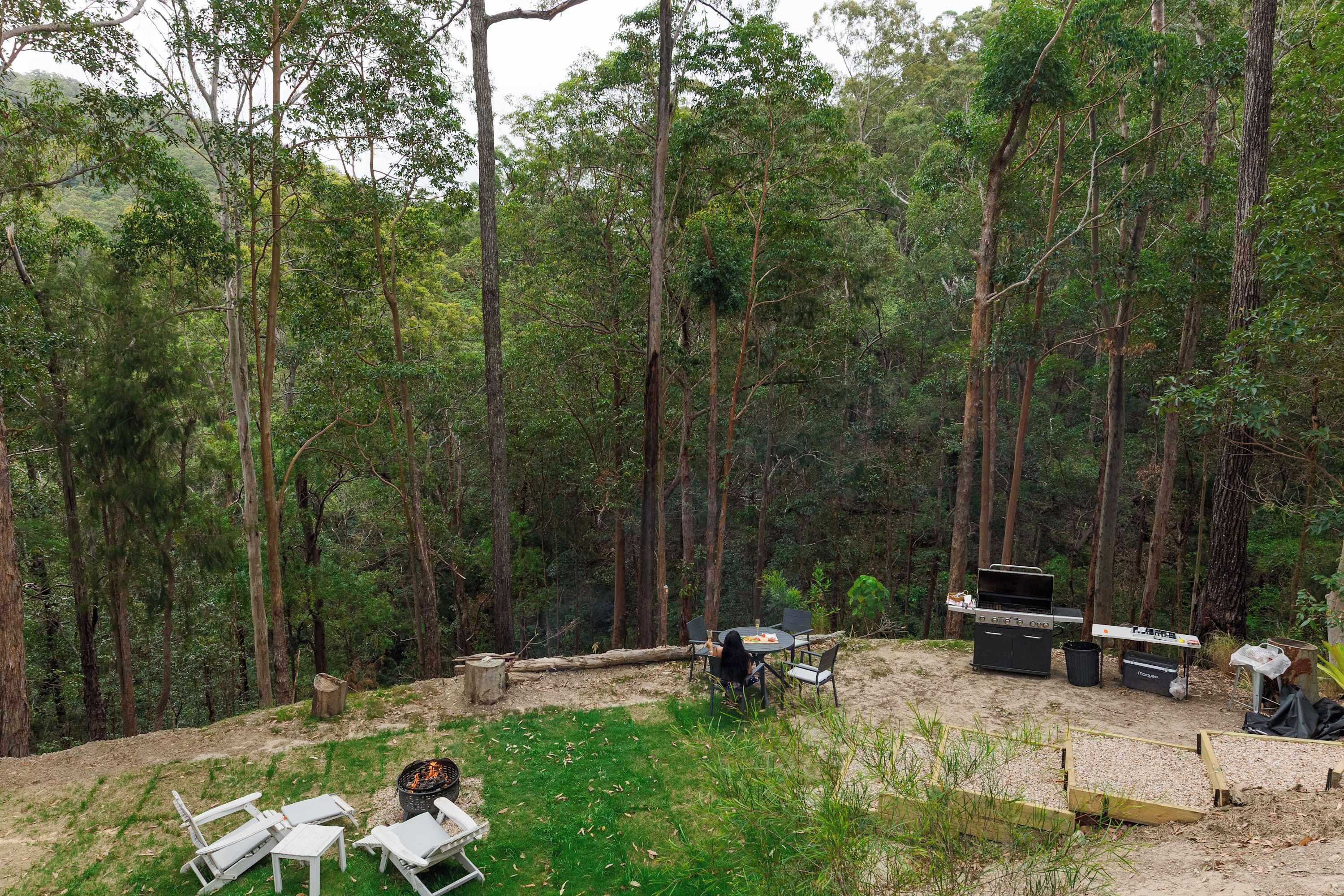 A forested campsite with a barbecue grill, seating area, and a fire pit.