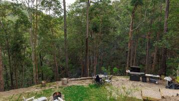 A forested campsite with a barbecue grill, seating area, and a fire pit.
