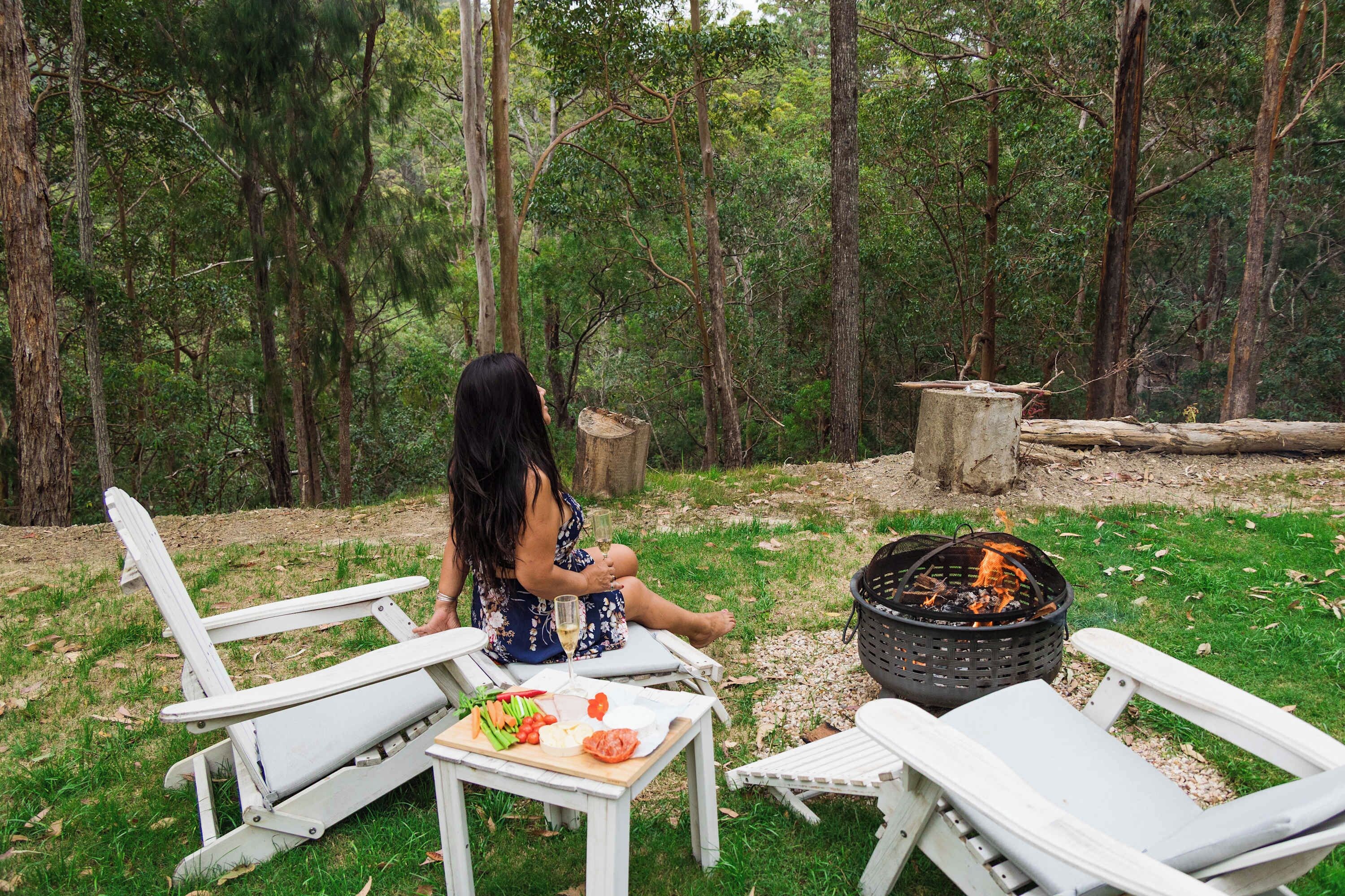 A woman sitting on a white chair outdoors with a fire pit and a table with food in front of her.