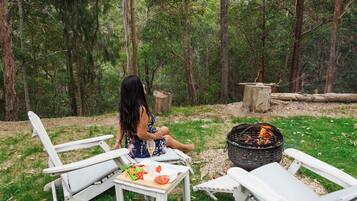 A woman sitting on a white chair outdoors with a fire pit and a table with food in front of her.