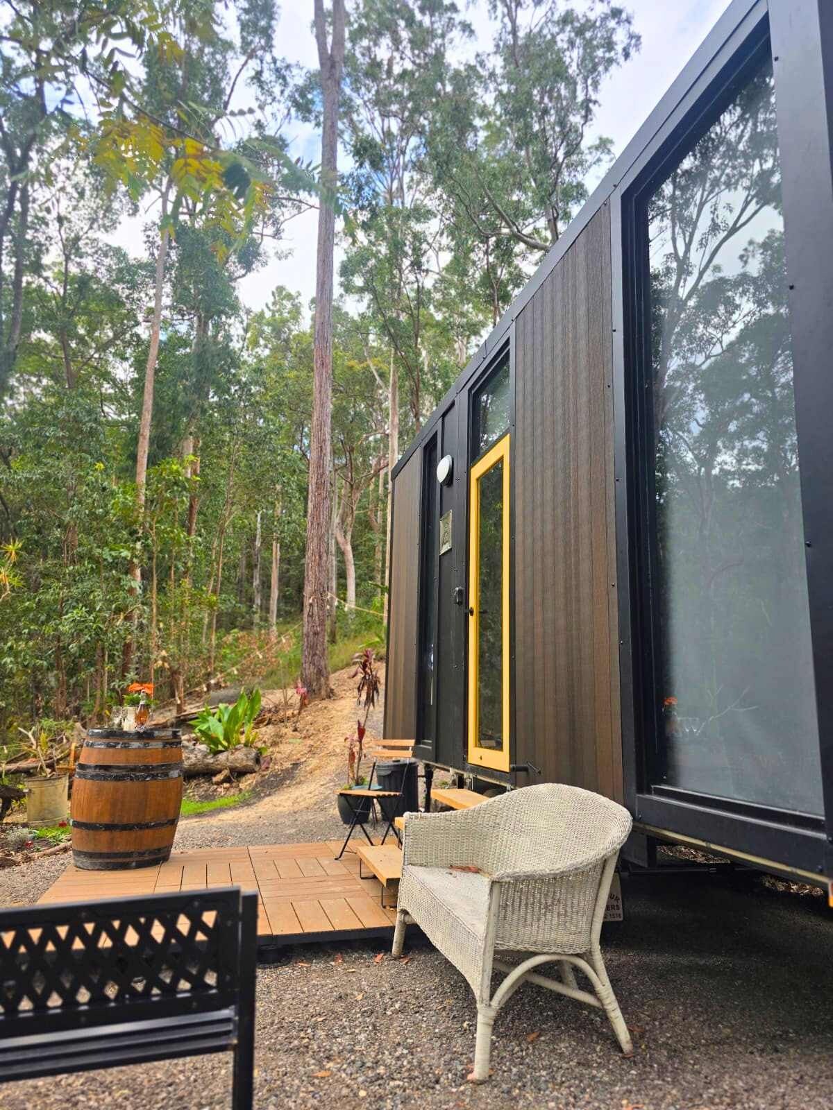 Une cabane moderne dotée d’une grande porte vitrée, entourée d’une zone boisée et agrémentée d’une terrasse en bois et de mobilier d’extérieur.