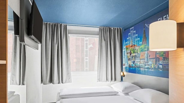 A hotel room with a bed, curtains, a window, a wall-mounted TV, and a lamp. - Ibis Budget Flensburg City (Flensburg)