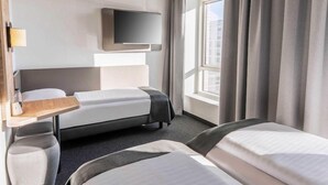 A hotel room with a bed, a TV mounted on the wall, a desk with a lamp, and a window with curtains. - Ibis Budget Flensburg City (Flensburg)