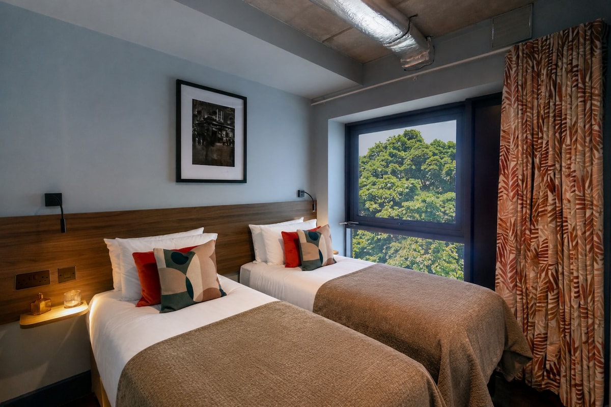 Une chambre d’hôtel avec deux lits, un tableau encadré au mur et une fenêtre donnant sur des arbres.
