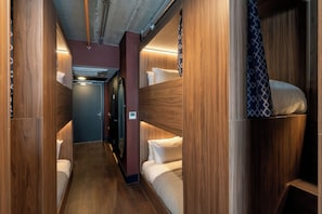 Un couloir étroit, avec des lits superposés de chaque côté, une porte au fond et un luminaire au plafond.