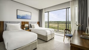 Habitación de hotel con dos camas, un escritorio, una silla, un ventanal con vistas y un cuadro en la pared.