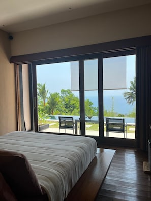 Un dormitorio con una cama grande, vistas a una piscina y palmeras, y un balcón con sillas.
