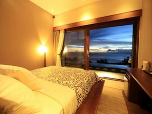 Habitación de hotel con cama, mesita de noche, lámpara y vista al océano al atardecer.