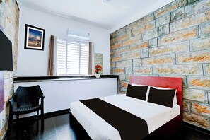 Hotellihuone, jossa on tiilikuviollinen seinä, punaisella päätyosalla varustettu sänky, sälekaihtimilla varustettu ikkuna, tuoli ja seinällä oleva kehystetty kuva.
