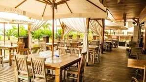 An outdoor restaurant with wooden tables and chairs, covered by large umbrellas, and a wooden deck. - Life Resort - Beira do Lago em Brasilia (Brasília)