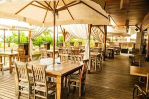 En udendørs restaurant med træborde og stole under store parasoller, samt en træterrasse.