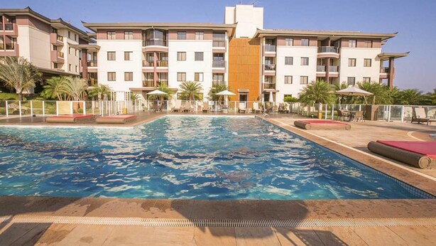 A swimming pool with a surrounding deck, a multi-story building with balconies, and palm trees. - Life Resort - Beira do Lago em Brasilia (Brasília)