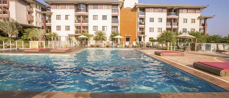 A swimming pool with a surrounding deck, a multi-story building with balconies, and palm trees.