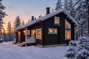 A dark wooden cabin with a porch and multiple windows, surrounded by snow-covered trees. - Kaakkoishovi by Aavalevi (Levi)
