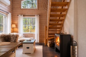 A cozy cabin interior with a wooden staircase, a fireplace, a sofa, and a window overlooking a forest. - Kaakkoishovi by Aavalevi (Levi)