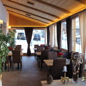 Restaurante com vigas de madeira no teto, grandes janelas com cortinas e mesas com cadeiras.