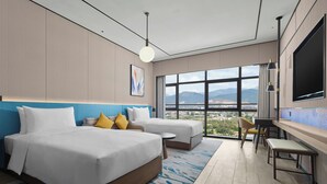 Habitación de hotel con dos camas, televisión, escritorio y ventana grande con vista a la ciudad.