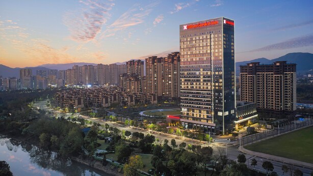 Un hotel moderno de gran altura con un logotipo rojo llamativo, rodeado de otros edificios y una masa de agua.