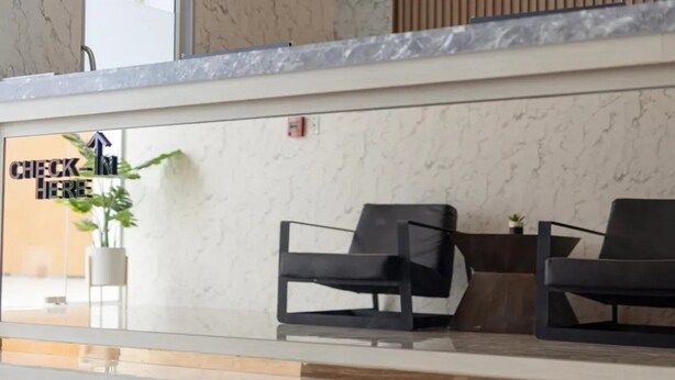 A hotel reception area with a marble counter, a sign that reads 'CHECK HERE', and two black chairs with a small table. - B2 Map Ta Phut Boutique & Budget Hotel (Rayong)