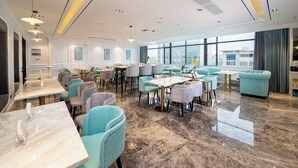 Un hall d’hôtel spacieux, doté de plusieurs espaces de détente, notamment des tables rondes et des canapés, ainsi que de grandes fenêtres offrant une vue sur la ville.