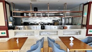 Un restaurante con cocina central, varias mesas con sillas y piso de baldosas.