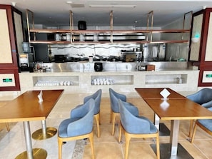 Restaurante com cozinha central, várias mesas com cadeiras e piso ladrilhado.