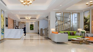 Um hall amplo com balcão de recepção, um lustre grande e área de estar com sofá e poltronas.