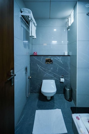 A compact bathroom with a toilet, a towel rack, a trash bin, and a small shelf with toiletries. - Zip By Spree Hotels Purple Orchid (Bengaluru)