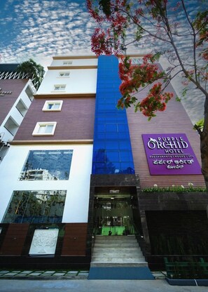 Un hotel con un letrero morado, fachada de vidrio azul y un árbol con flores rojas.
