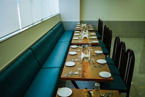 A dining area with a long table set for multiple guests, a green cushioned bench, and chairs arranged around the table. - Zip By Spree Hotels Purple Orchid (Bengaluru)