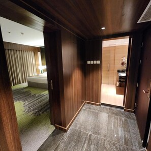 Una habitación de hotel con puerta de madera, un baño con inodoro y lavamanos, y una sala de estar con sofá y cortinas.