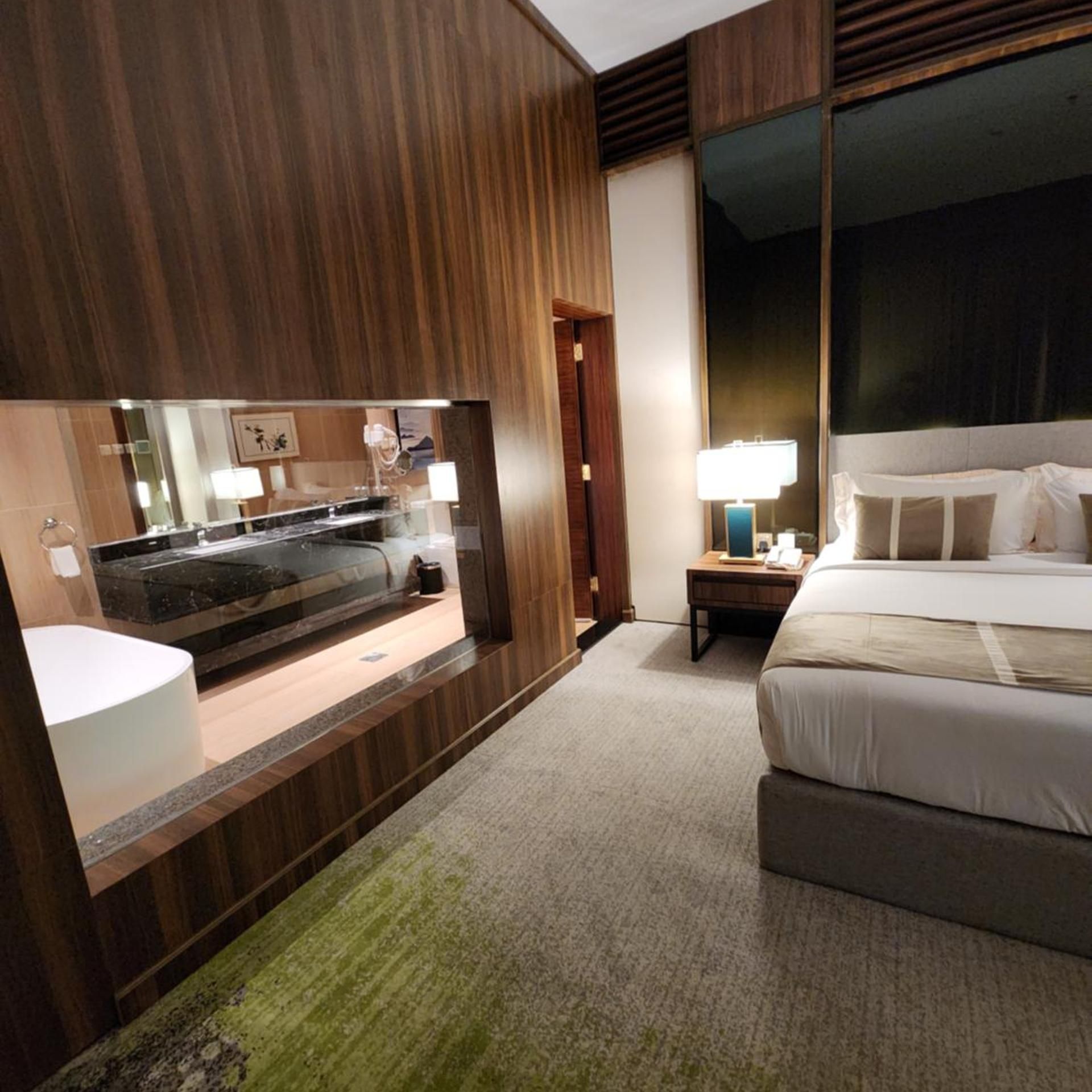 A hotel room with a large bed, a bedside table with a lamp, a mirror, and a view of the outside.