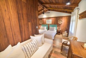 A wooden-floored room with a sofa, a small table, and a chair. There are two beds with green headboards and white bedding.