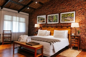 A hotel room with a brick wall, a wooden bed, two bedside tables with lamps, a bench, and a window with sheer curtains.
